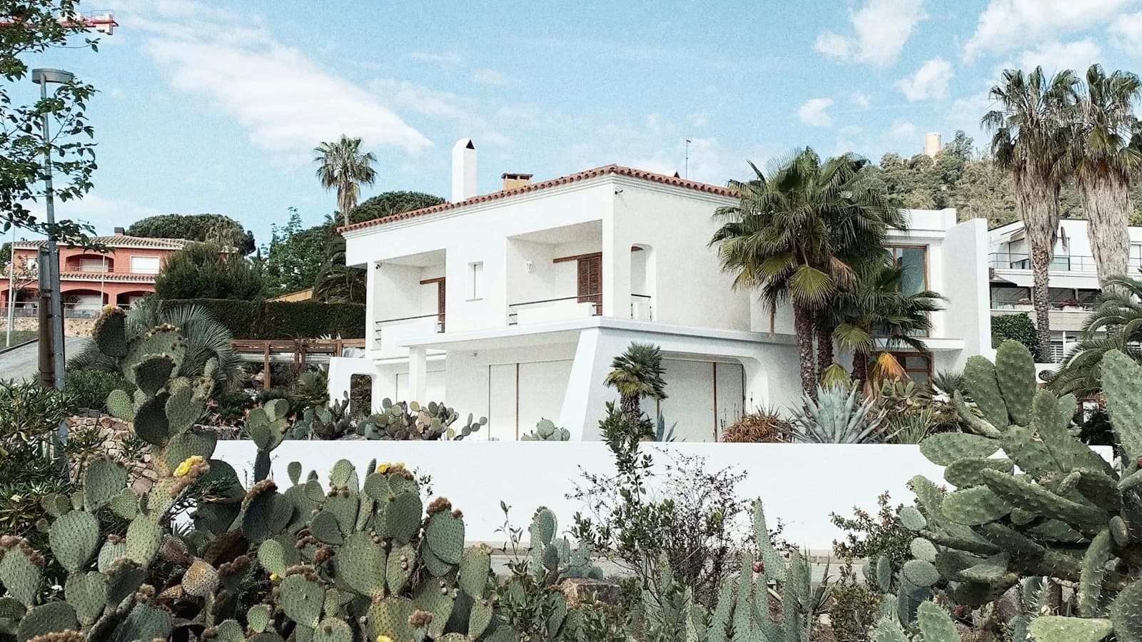 A white two-storey house with a tiled roof, surrounded by cactus, palm trees and garden plants under a blue sky