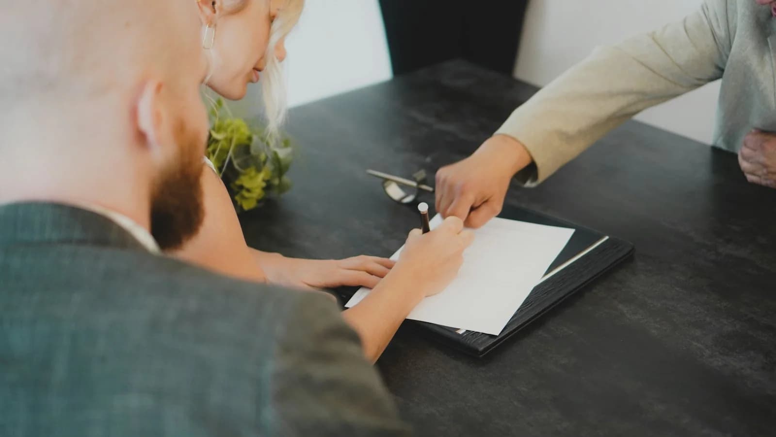 Two buyers seated at a desk signing a document while an adviser points to where to sign